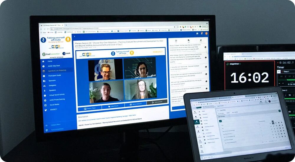 Desktop monitor on a dark desk showing a virtual event platform, with a blue navigation sidebar, a plenary session video grid of four speakers and a chat panel to the right, and an open laptop beside it