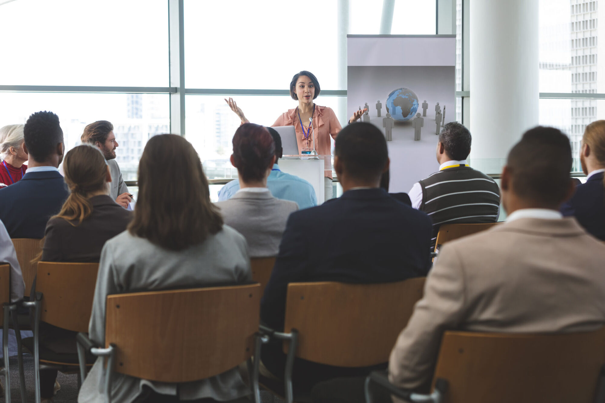 Woman in a pink blouse speaks from a lectern to a seated audience in a bright glass-walled conference room