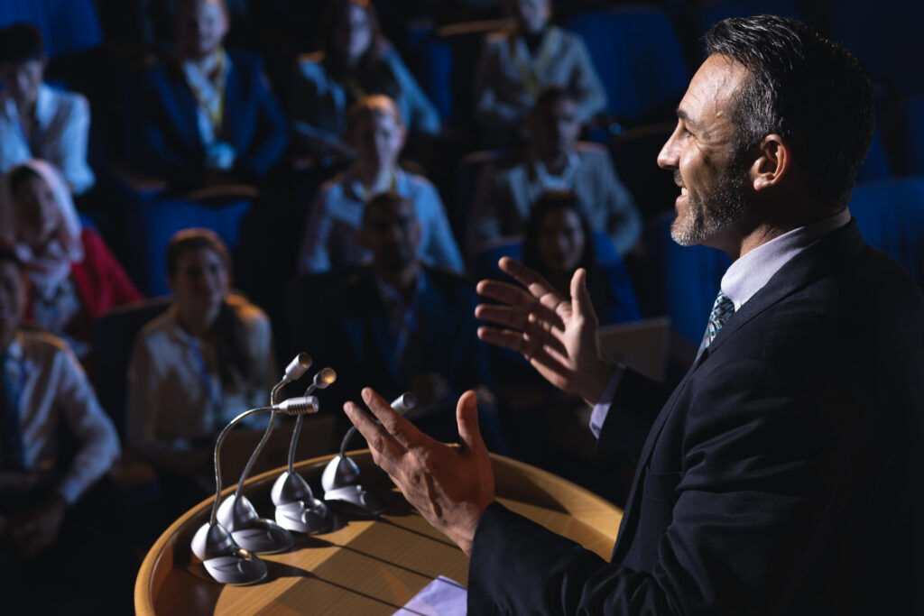 Bearded man in a dark suit gestures with both hands as he speaks at a lectern of microphones to a dimly lit seated audience