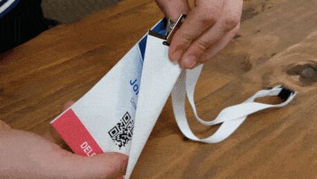 Hands twisting a Global Conference name badge printed for John Smith, with a QR code and fabric lanyard, to show it does not tear
