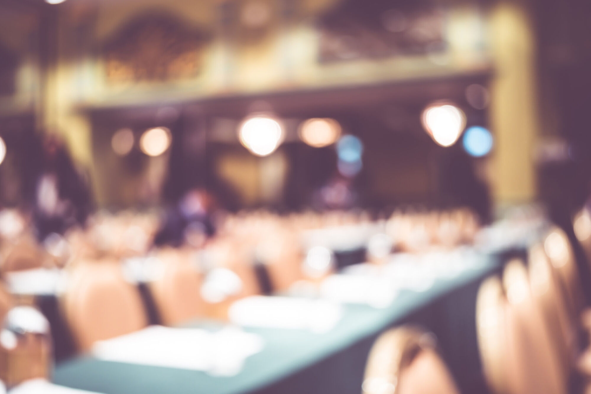Deliberately out-of-focus view of a banquet hall laid with round tables and glassware in front of a lit stage