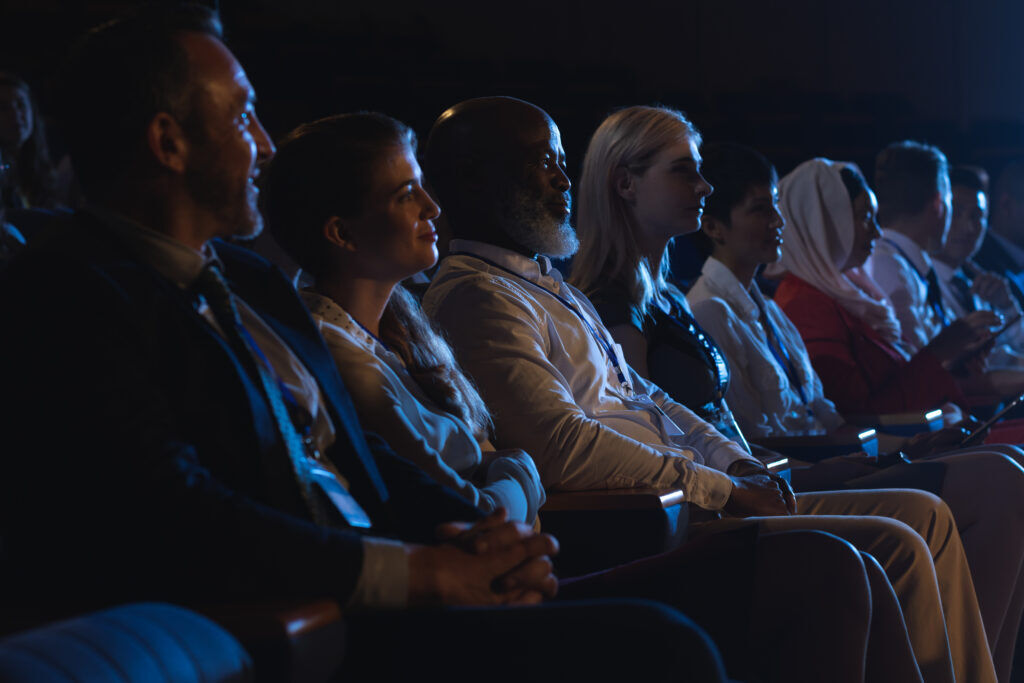 Row of smartly dressed attendees wearing lanyards, seated in a darkened auditorium watching a presentation
