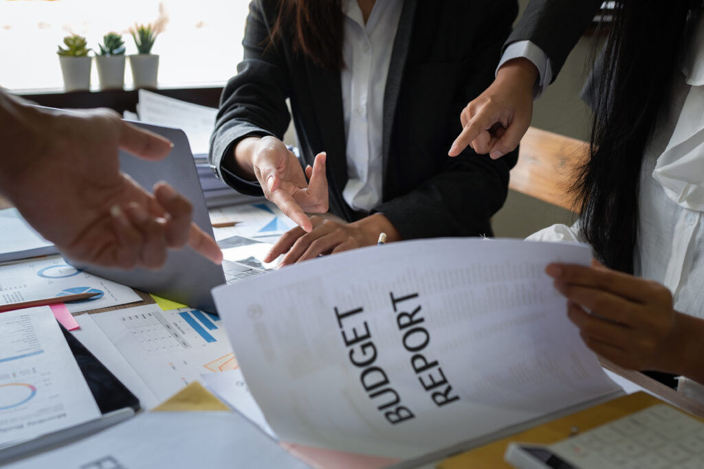 Colleagues gathered over a desk of charts and spreadsheets, one holding a document headed Budget Report