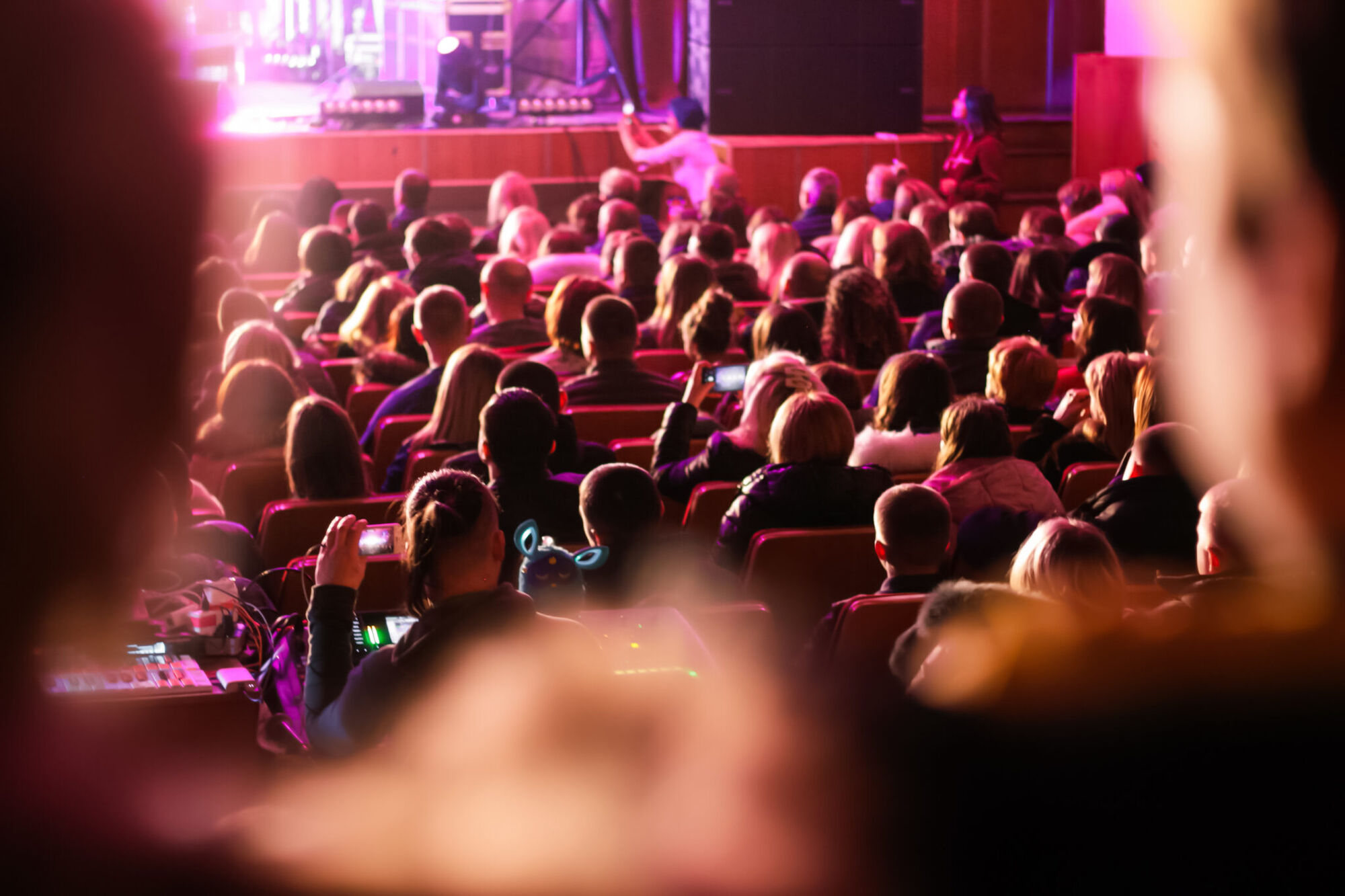 Packed theatre audience seen from behind under pink stage lighting, several people filming the stage on cameras and phones