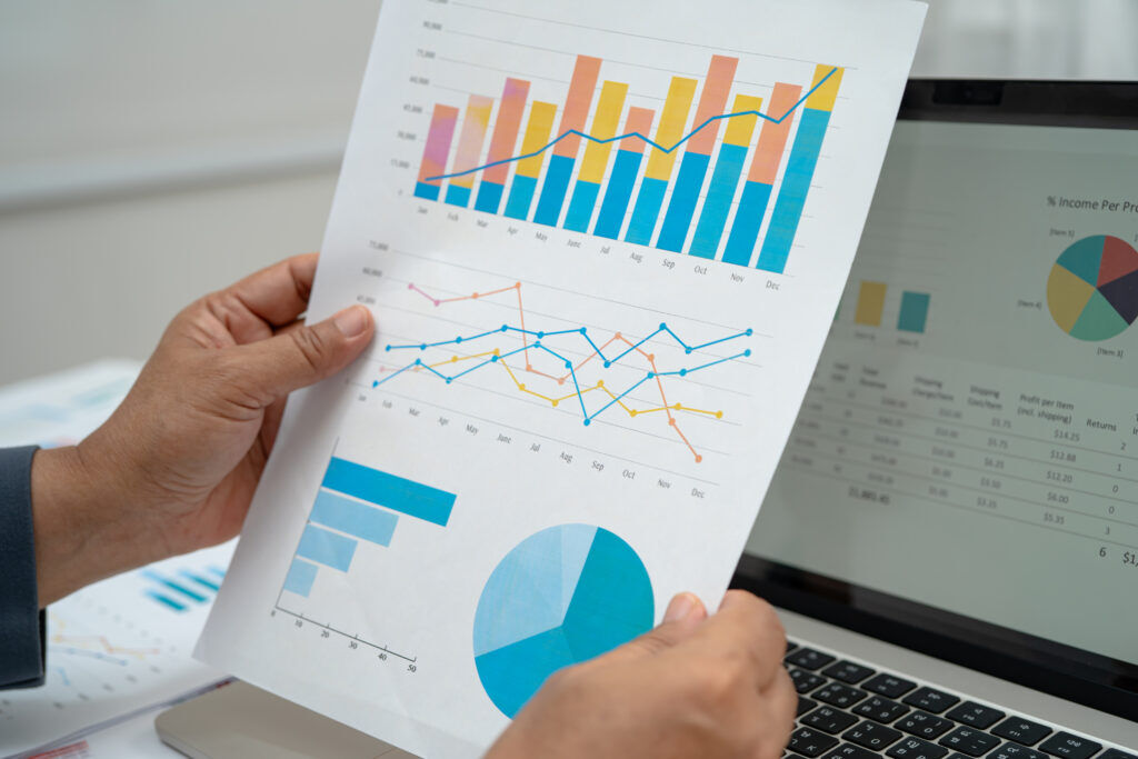 Hand holding a printed page of bar, line and pie charts in front of a laptop displaying a revenue spreadsheet