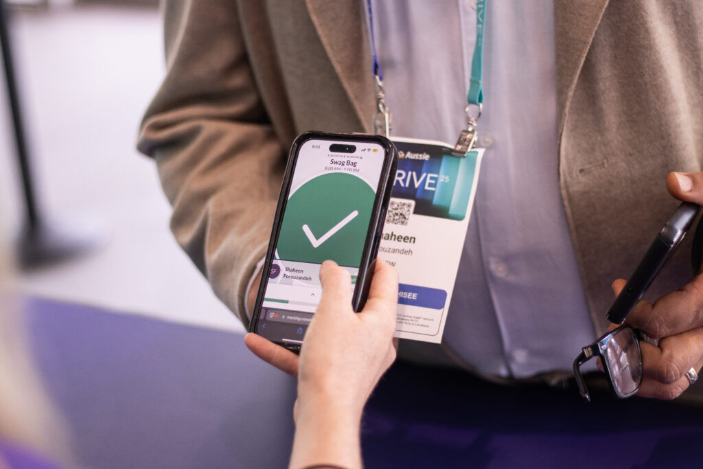 Person holding a phone showing a large green tick confirming Swag Bag collection, next to an attendee wearing a conference badge on a lanyard