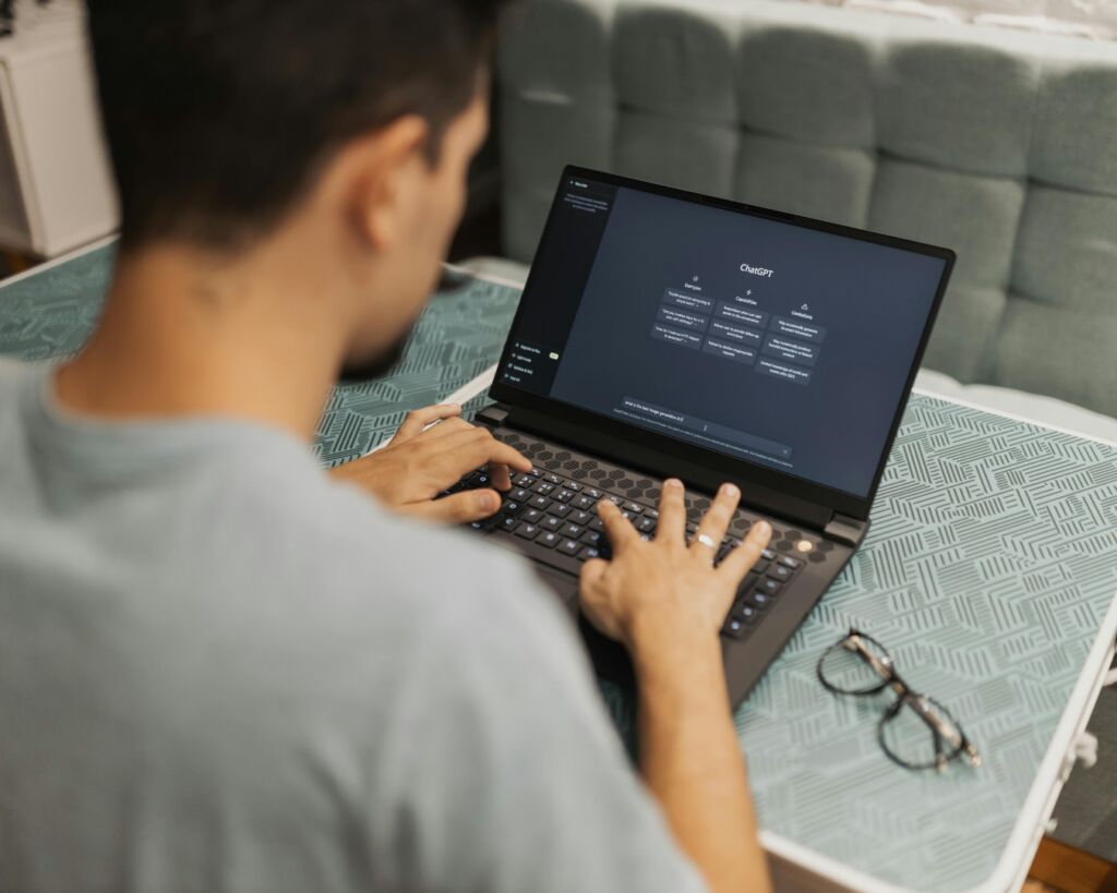 Man seated at a low table typing on a laptop that displays the ChatGPT home screen with example prompts, glasses beside him