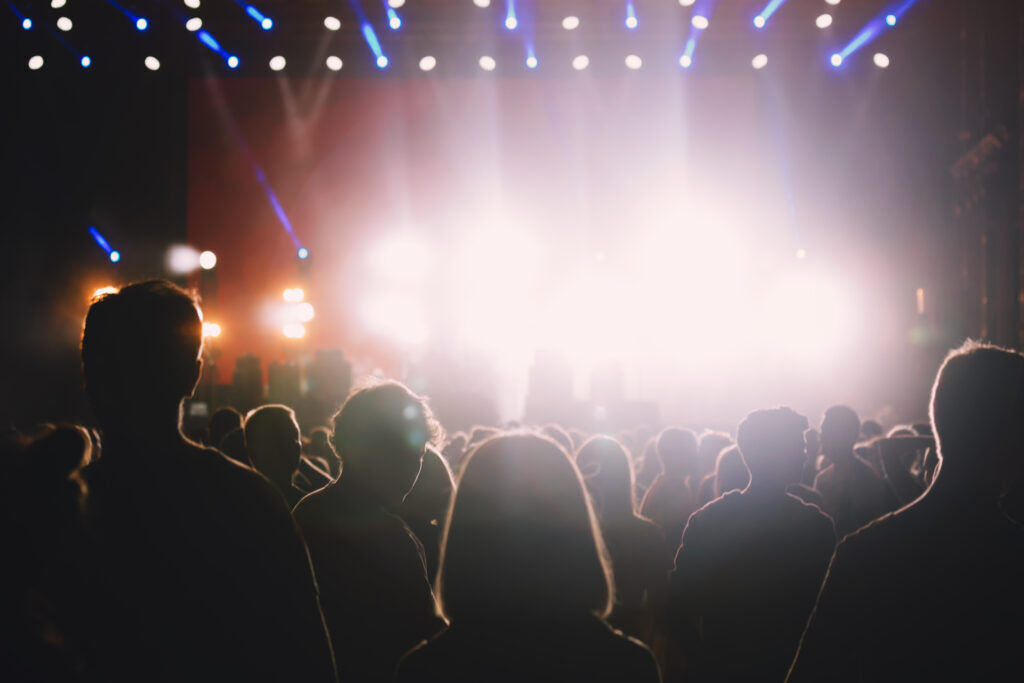 Silhouetted crowd standing in front of a brightly lit festival stage with blue spotlights overhead