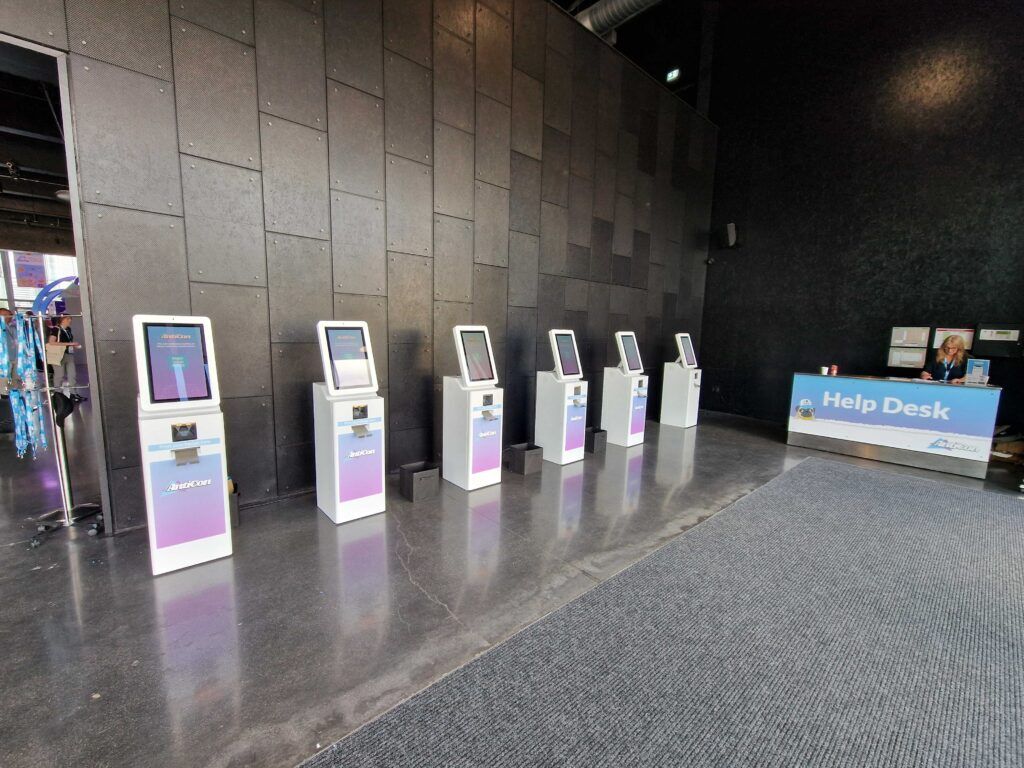 Six white self-service check-in kiosks branded AntiCon lined up along a dark panelled wall beside a staffed Help Desk