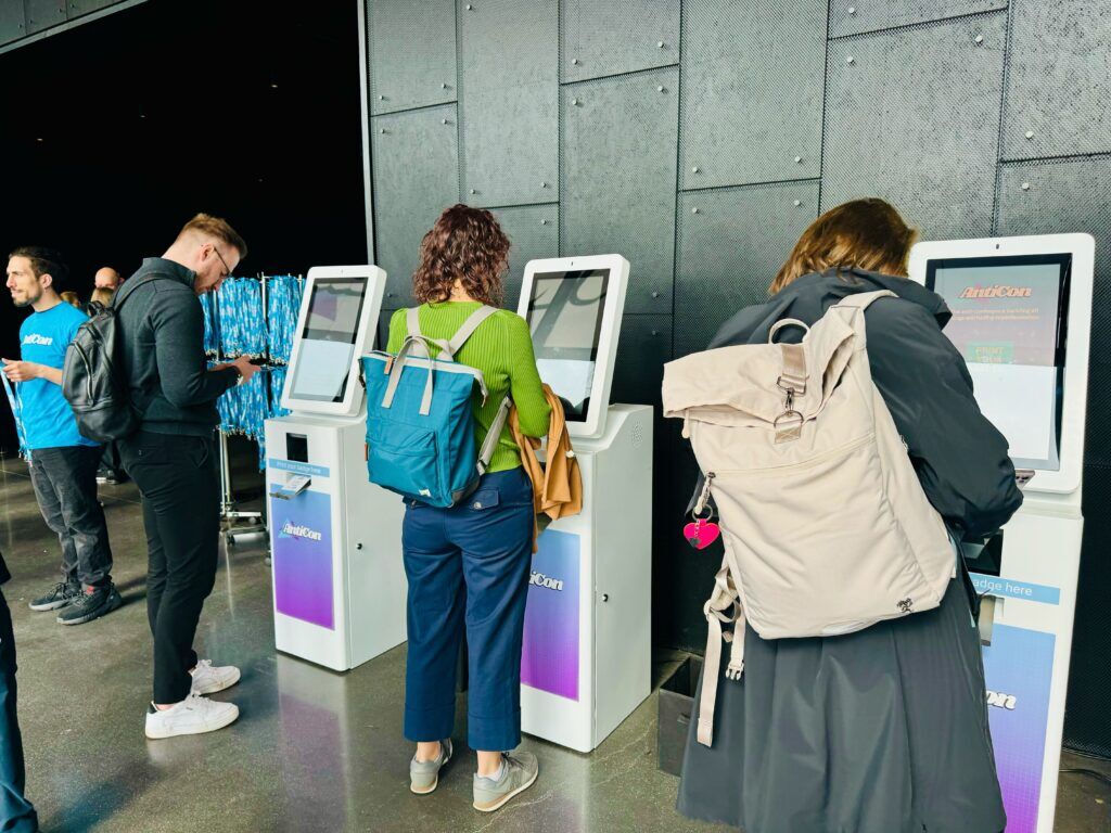 Three attendees with backpacks using AntiCon-branded touchscreen check-in kiosks at a venue, with staff and a lanyard rail behind them