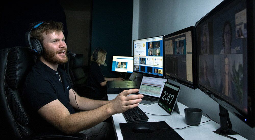 Man in a headset at a broadcast desk of monitors showing a virtual session and speaker video panels, with a countdown timer reading 2:47