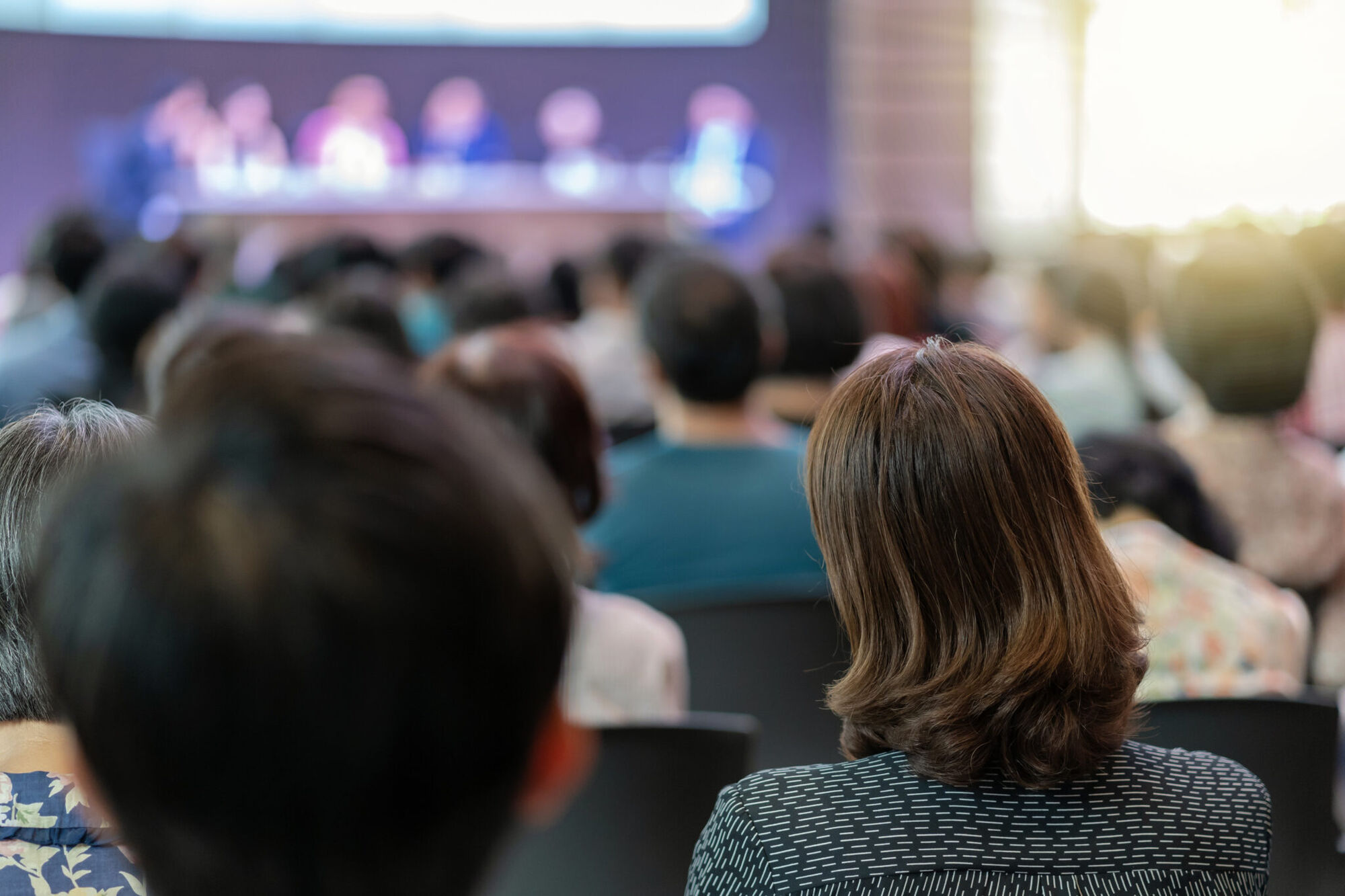 Conference audience seen from behind, facing a blurred row of panel speakers seated at a table on stage