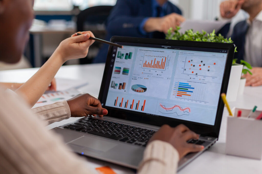 Colleagues at a desk looking at a laptop dashboard of bar, pie and line charts, one pointing at the screen with a pen