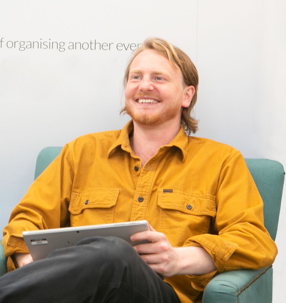 Smiling bearded man in a mustard shirt sitting in a teal armchair with a laptop on his knee, wall text partly visible behind him