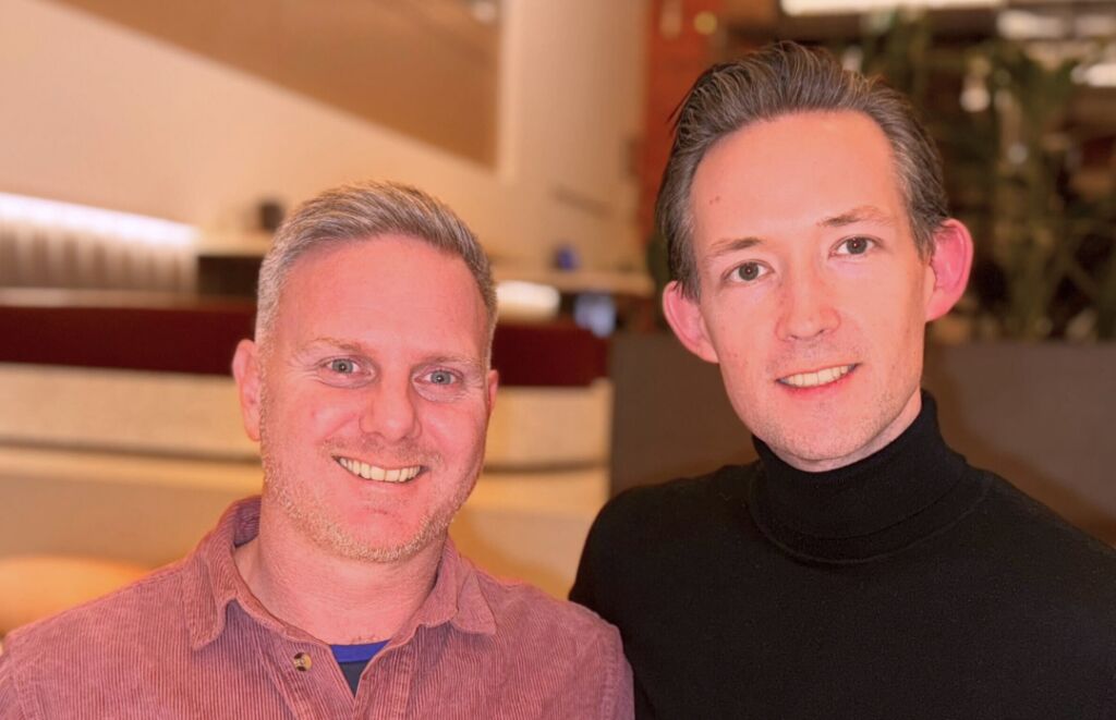 Two men smiling side by side for a close-up photo in a bright hotel lobby, one in a pink corduroy shirt and one in a black polo neck