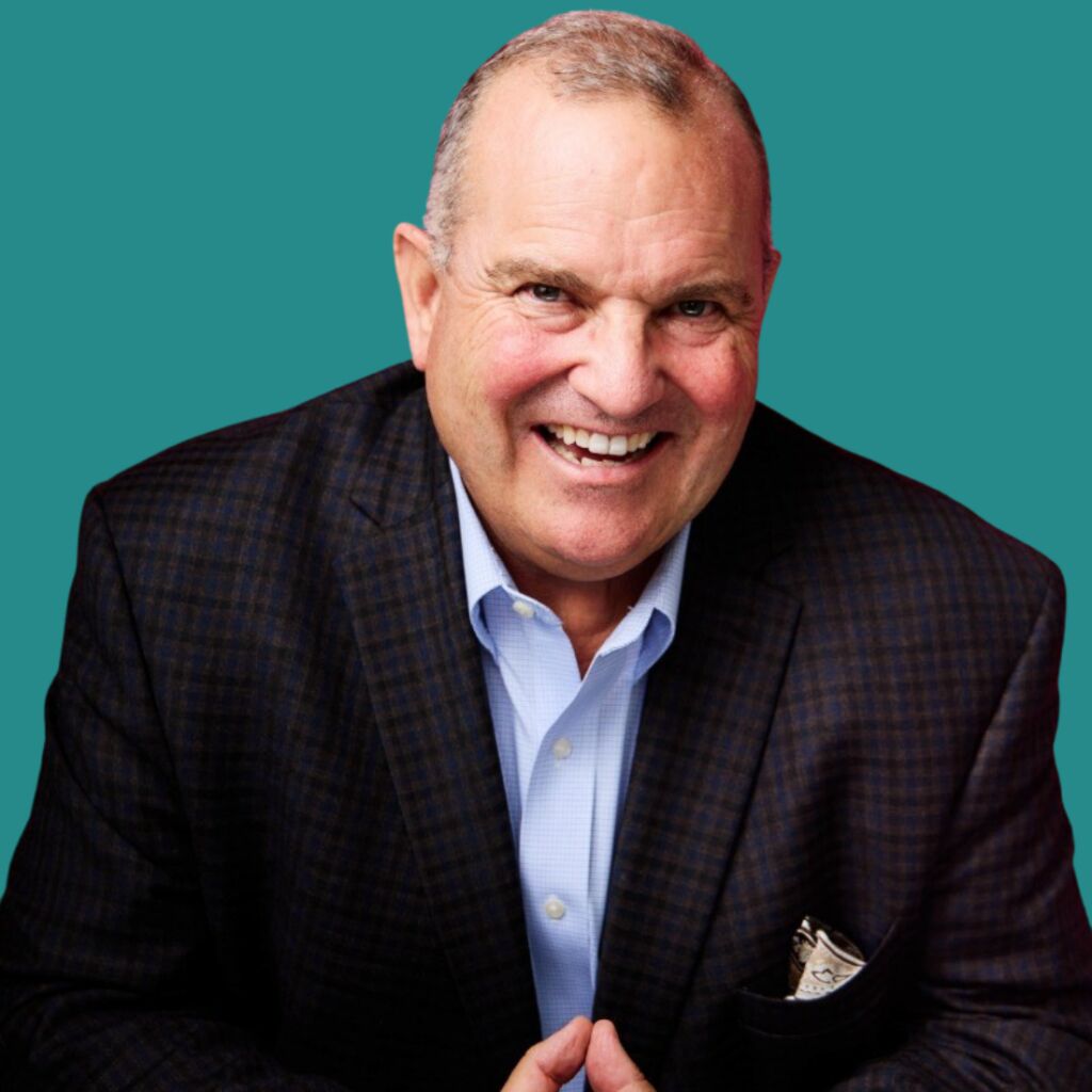 Smiling man in a dark checked blazer and pale blue shirt, hands together, photographed against a plain teal background