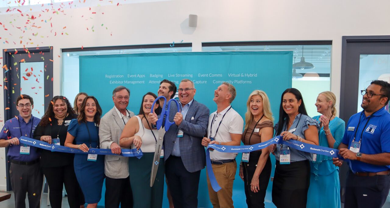 Group of colleagues and guests cutting a blue Gwinnett Chamber ribbon with giant scissors amid falling confetti, in front of a teal CrowdComms services backdrop
