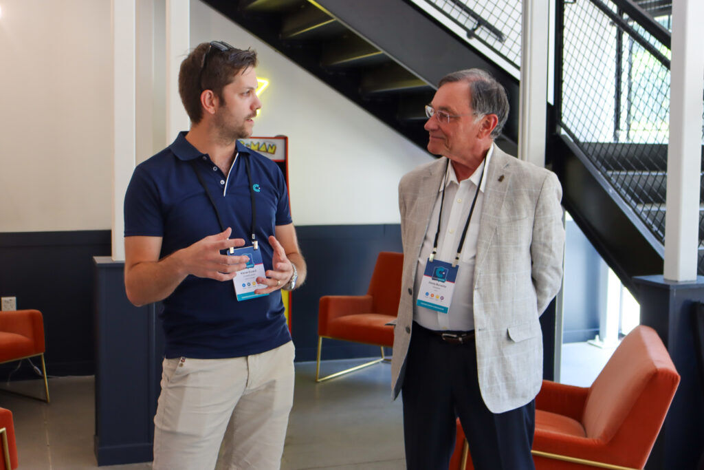 Man in a navy CrowdComms polo shirt talking with an older guest in a grey blazer, beside orange armchairs and an arcade machine under a staircase