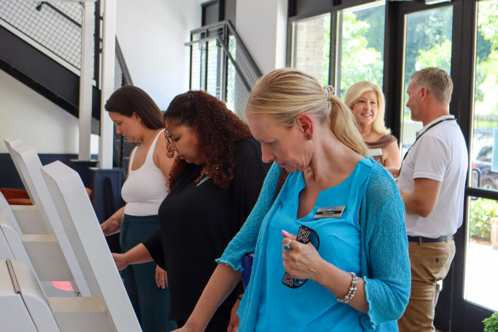 Three guests using white self-service check-in kiosks in a sunlit office, with other visitors talking by the windows behind