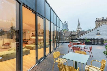 Rooftop terrace with yellow and orange cafe tables beside a glass-walled lounge, with London rooftops and a church spire beyond