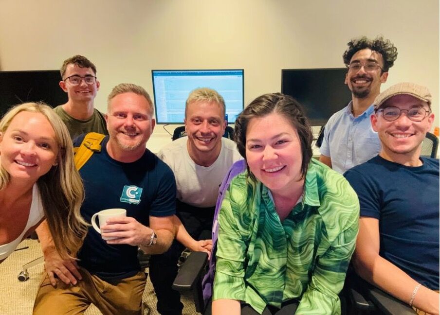 Seven colleagues leaning in together and smiling in an office, one in a CrowdComms T-shirt, with desk monitors behind them