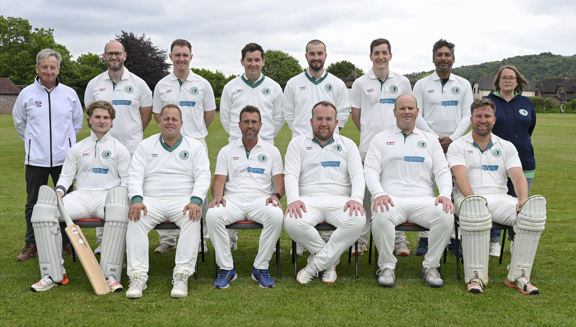Cricket team photographed in two rows on a village ground, wearing whites with crowdcomms sponsor logos across the chest