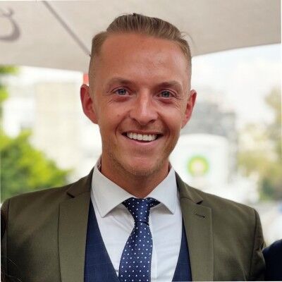 Smiling man in an olive suit with a navy waistcoat and blue polka-dot tie, photographed outdoors under a marquee