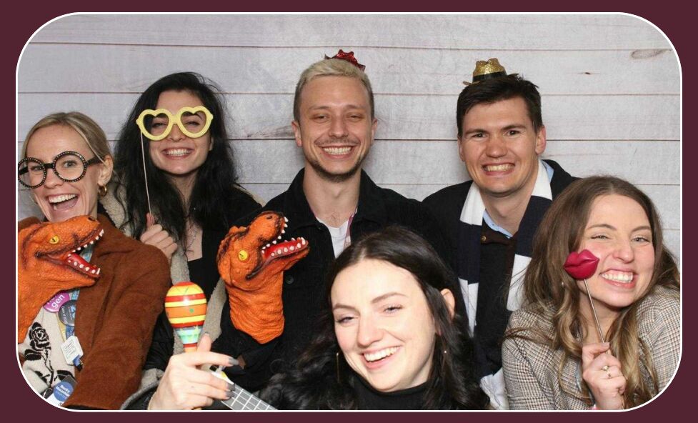 Photo booth shot of six people laughing with props including oversized glasses, dinosaur hand puppets, maracas and lips on a stick