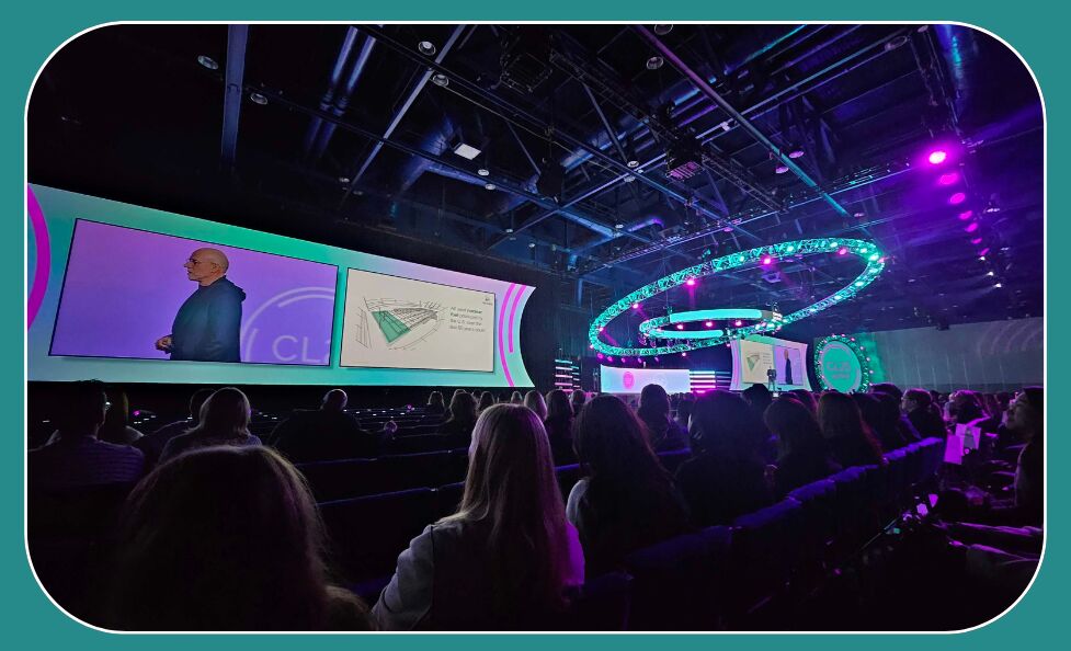 Audience in a darkened conference hall watching a speaker on twin screens, beside a teal and pink lit stage with a curved lighting rig