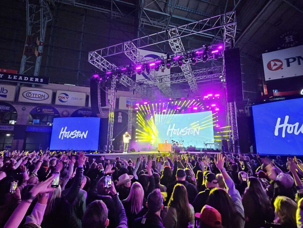 Crowd with hands raised at an indoor concert, three screens reading "Houston" behind a performer on a brightly lit stage