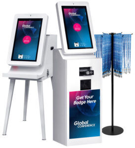 Two white self-service check-in kiosks, one with a built-in badge printer labelled "Get Your Badge Here", next to a rotating lanyard stand
