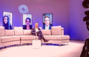 Two people seated on a curved sofa on a purple-lit studio set, with three portrait screens on the wall behind them