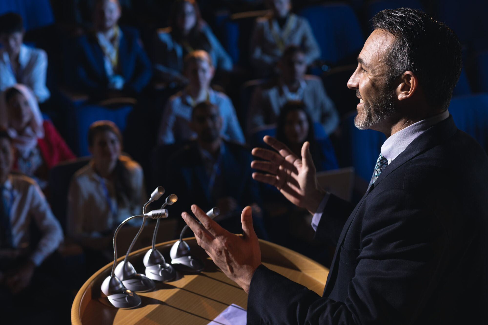 Bearded man in a dark suit gestures with both hands as he speaks at a lectern of microphones to a seated audience in a dim auditorium