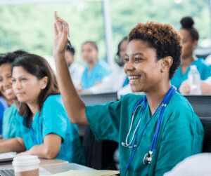 Smiling delegate in teal scrubs with a stethoscope in a lecture room, while a colleague behind her raises a hand to ask a question
