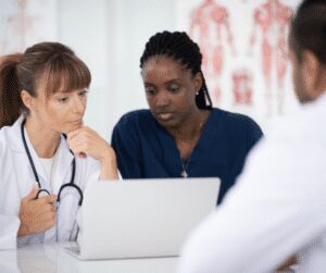 Two clinicians in a white coat and scrubs studying a laptop together, with anatomy posters on the wall behind
