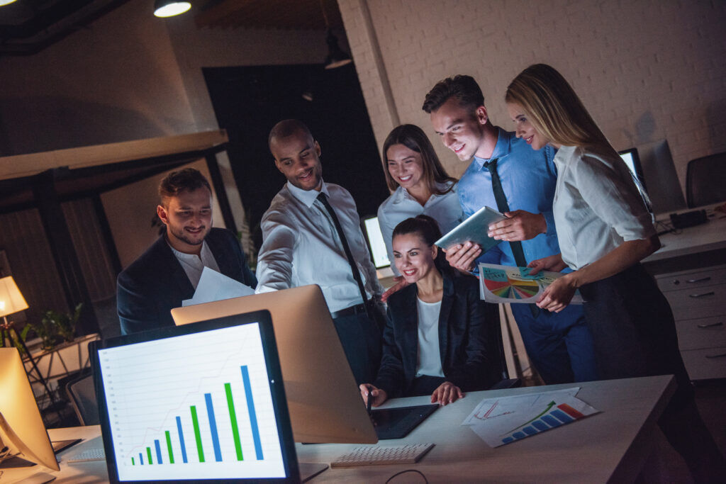 Six colleagues in office wear gathered around a desktop monitor at night, smiling at a rising bar chart, with printed charts in hand