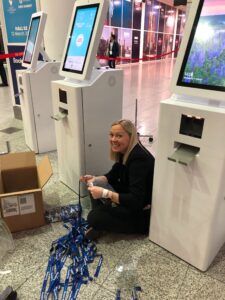 Woman crouched beside a row of badge kiosks in a venue, untangling a pile of blue lanyards next to an open box