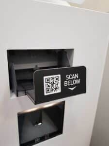 Close-up of a kiosk scanner hatch with a black label reading Scan below next to a QR code, above the badge dispensing slot