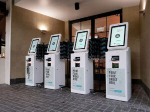 Four white self-service badge printing kiosks lined up in a venue entrance, each with a touchscreen and a Print your badge here panel