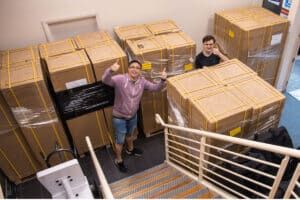 Two people give a thumbs up beside large shrink-wrapped crates of stacked kiosk equipment in a storage room