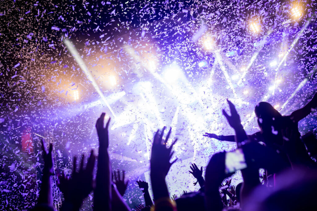 Crowd with hands in the air in front of a brightly lit stage, silver confetti falling through purple beams
