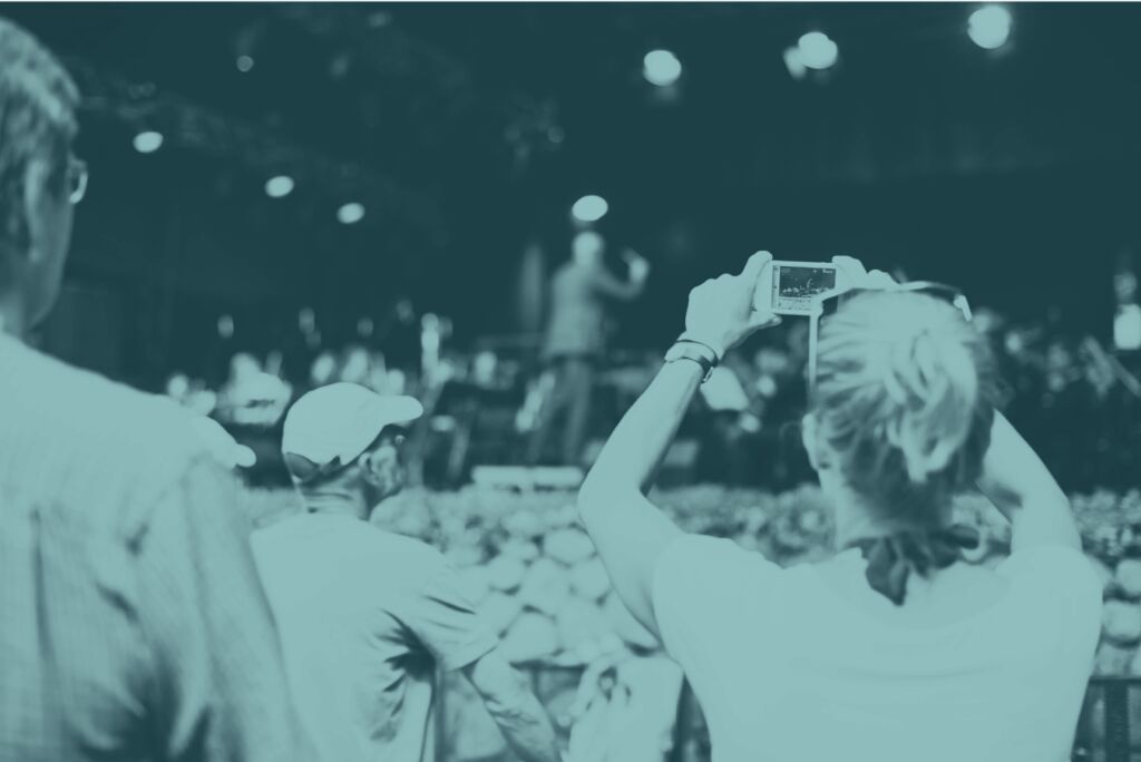 Teal-tinted photo of festival-goers watching a performer on stage, one woman holding a camera up to take a picture