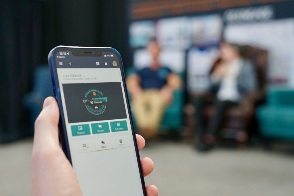 Hand holding a phone showing an event app live stream page with Popout, Stream and Schedule buttons, while two people are interviewed on stage behind