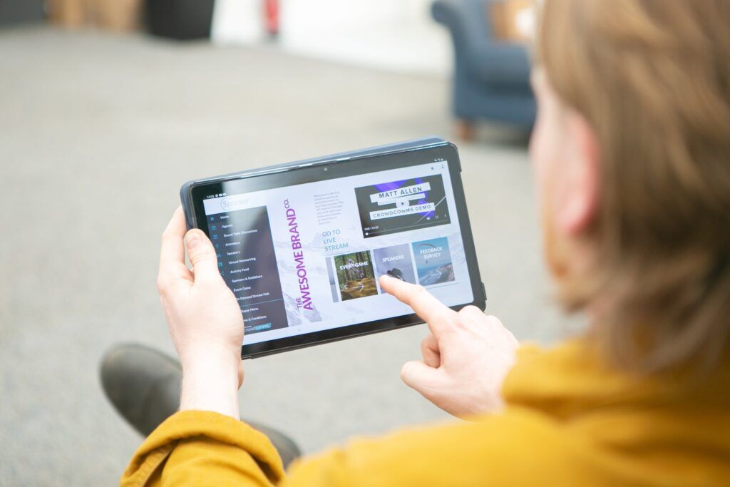 Over-the-shoulder view of a person tapping a tablet showing a branded event platform with a video of a speaker and tiles for Event Game, Speakers and Feedback Survey