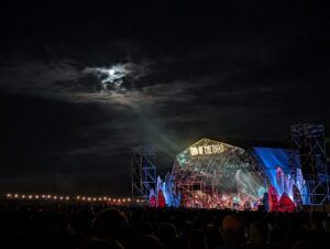 Outdoor festival stage lit up at night under a cloudy moonlit sky, with a crowd silhouetted in the foreground