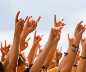 Many hands raised against a pale sky making rock horn gestures, some wearing festival wristbands