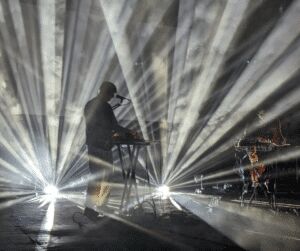 Silhouetted performer at a keyboard on a dark stage, surrounded by fanned-out white spotlight beams