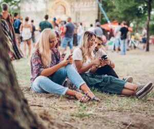 Three young festival-goers sitting on the grass under a tree looking at their phones, with crowds and stalls behind them