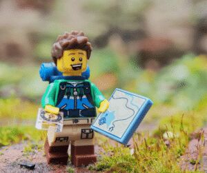 Close-up of a Lego figure dressed as a hiker with a backpack, holding an open map, on mossy ground
