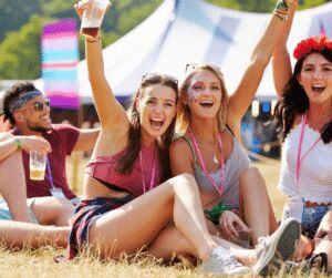 Four friends in festival outfits and glitter face paint sitting on the grass, laughing with arms raised, marquees behind them